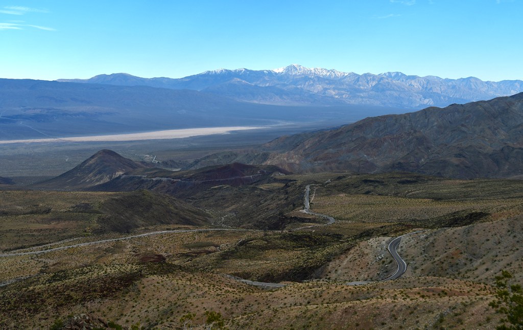 A panoramic view of a desert landscape with rolling hills and a winding road, framed by distant mountain ranges under a clear blue sky.