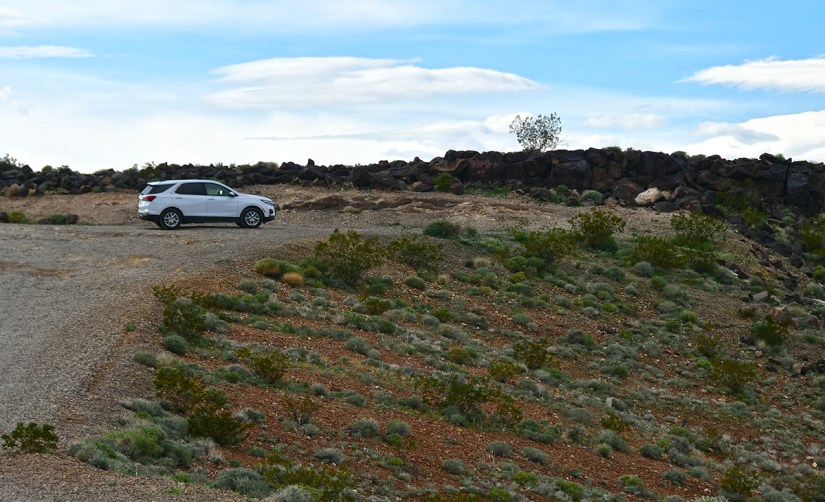 A white SUV parked on a gravel path surrounded by a hilly, green landscape with scattered shrubs and a rocky background under a partly cloudy sky.