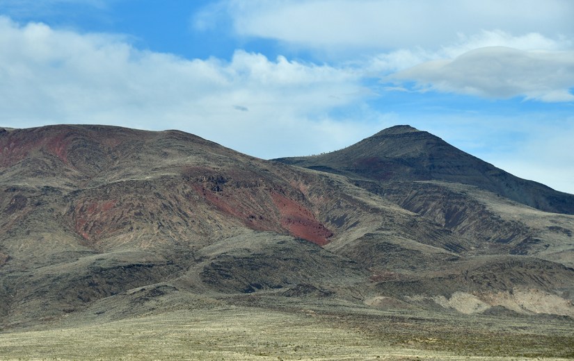 Dramatic mountain landscape with layered, rocky terrain and patches of red earth under a partly cloudy sky.