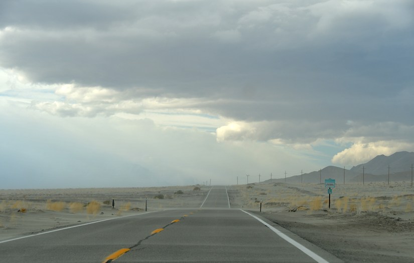 A long, straight road stretching into the distance under a cloudy sky, with mountains faintly visible on the horizon and a few roadside signs.
