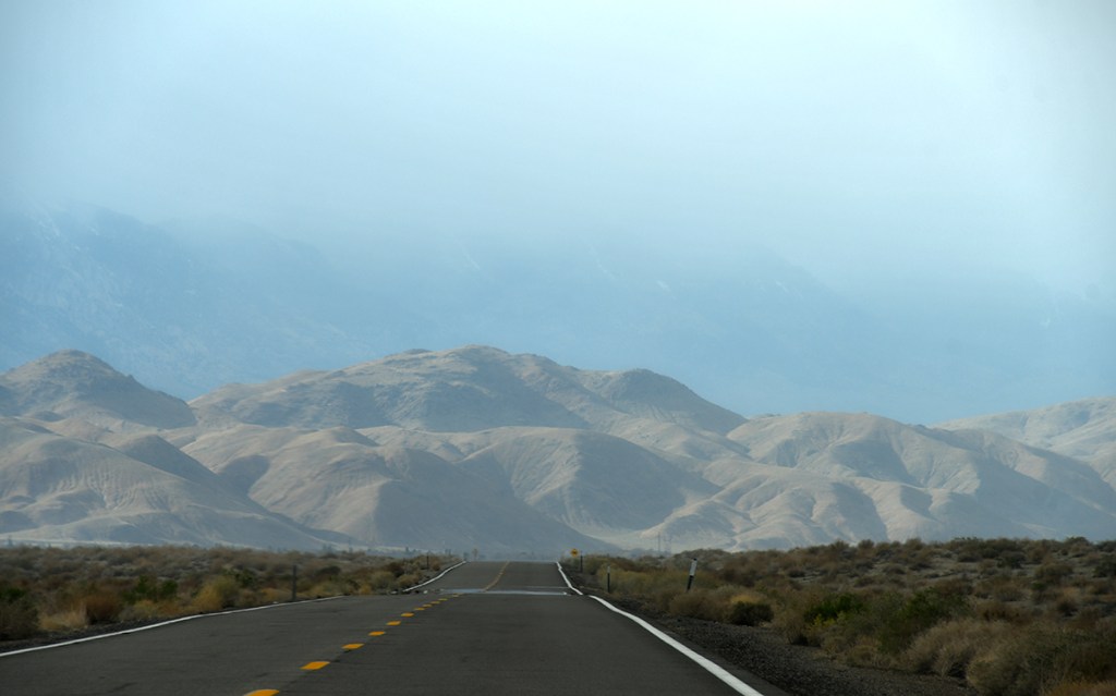 A long, empty road stretches through a desert landscape, leading towards hazy mountains in the background.