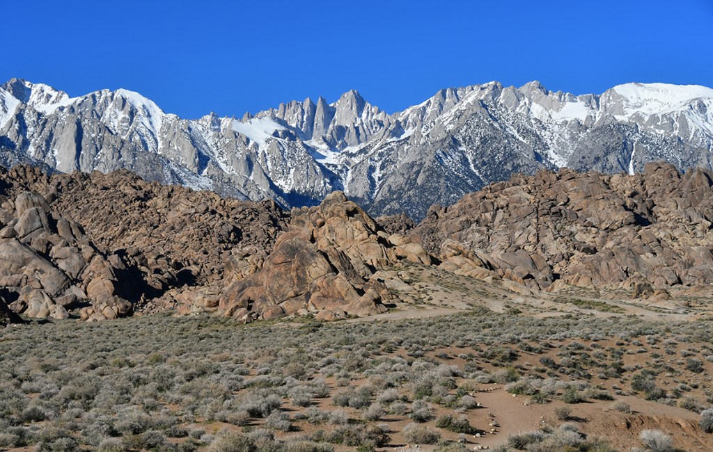 A panoramic view of rugged mountain peaks covered in snow, with rocky formations and sparse vegetation in the foreground.