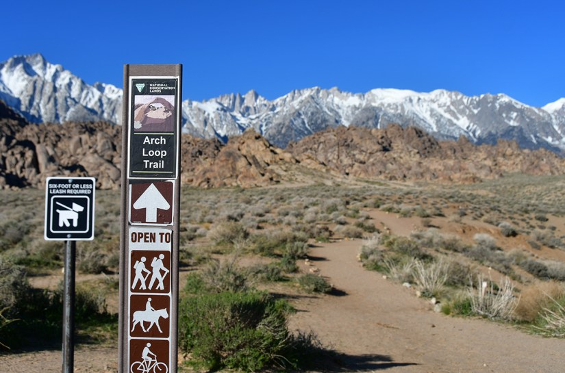 Signpost for Arch Loop Trail with directions for hikers, horseback riders, and cyclists, surrounded by rocky terrain and snow-capped mountains under a clear blue sky.