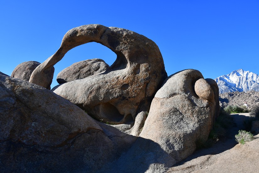 A scenic landscape featuring large, uniquely shaped rock formations under a clear blue sky, with snow-capped mountains in the background.