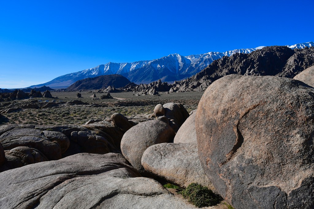 A panoramic view of rugged rocks in the foreground with a snow-capped mountain range in the background, under a clear blue sky.