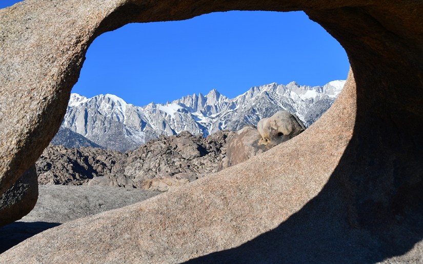 View through a rock archway framing snow-capped mountains against a clear blue sky.
