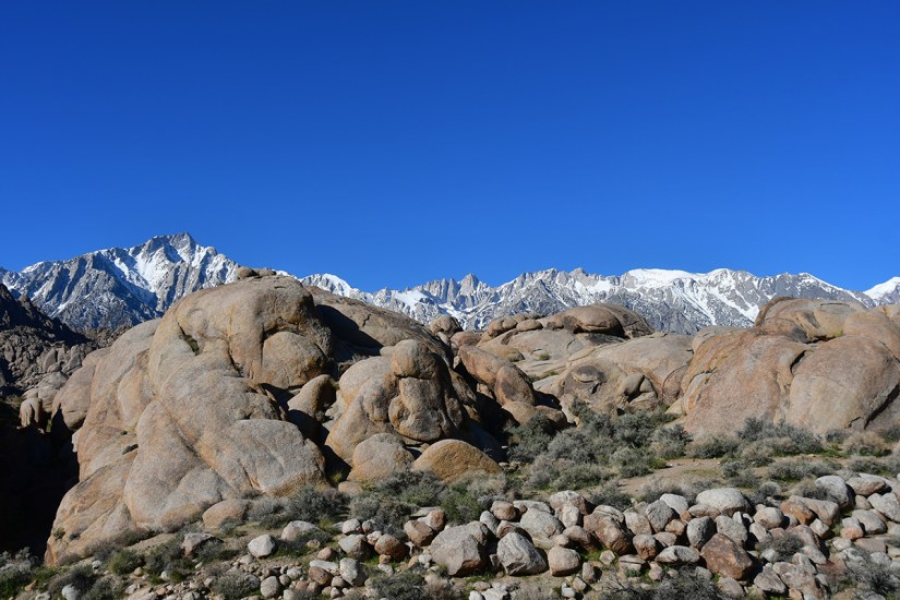 Rocky landscape with large boulders in the foreground and snow-capped mountains against a clear blue sky in the background.