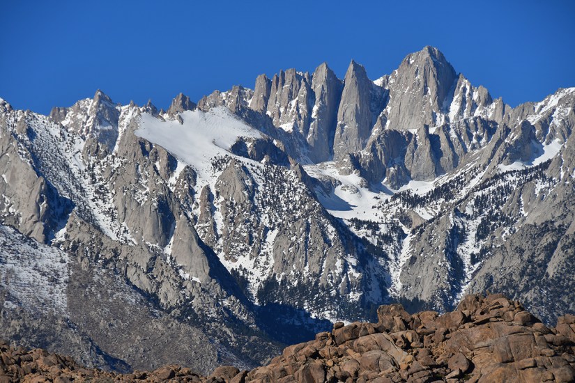 Snow-covered mountain peaks under a clear blue sky, with rocky terrain in the foreground.