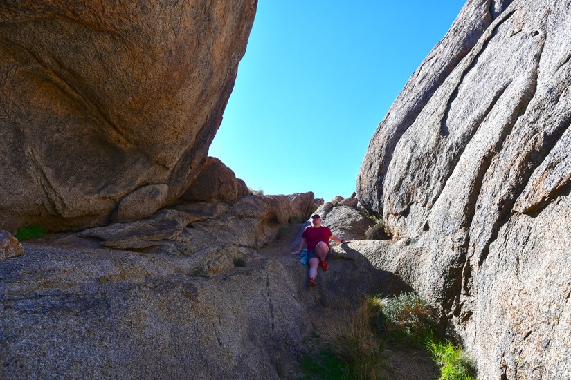 A person sitting on a rocky path between two towering rock formations under a clear blue sky.