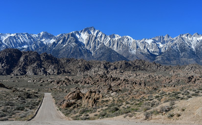 A dirt road winding through a rocky desert landscape with snow-capped mountains in the background under a clear blue sky.