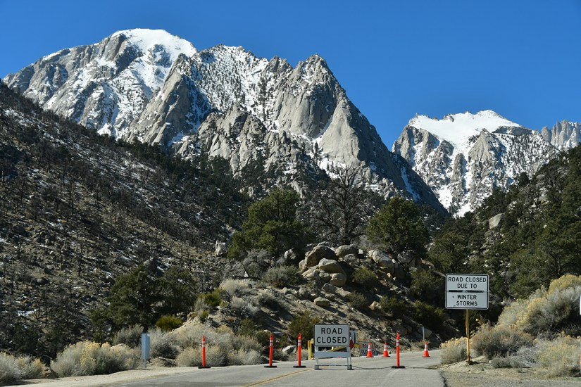 Snow-capped mountains with a closed road sign indicating winter storm conditions, surrounded by trees and rocky terrain.