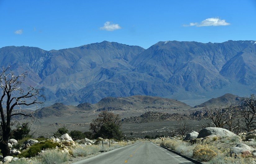 A scenic view of a road leading into a mountainous desert landscape, featuring dry terrain, large rocks, and sparse vegetation under a clear blue sky.