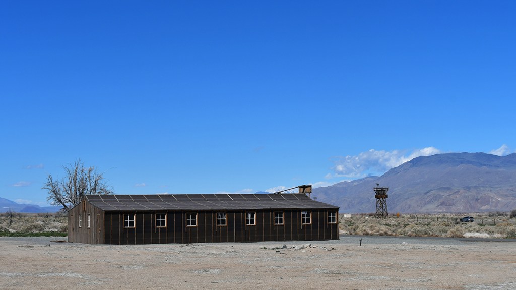 An old wooden building standing alone in a barren landscape beneath a clear blue sky, with distant mountains and a watchtower in the background.