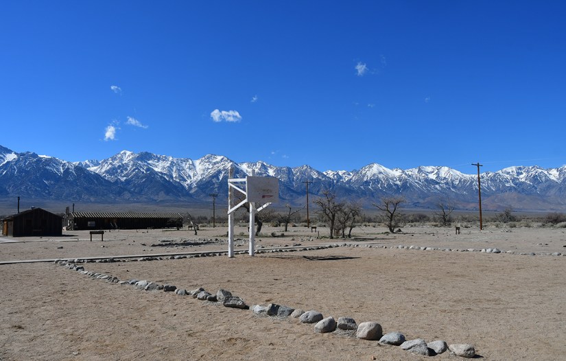 A basketball hoop stands in a dry landscape surrounded by a stone boundary, with snow-capped mountains and a clear blue sky in the background.