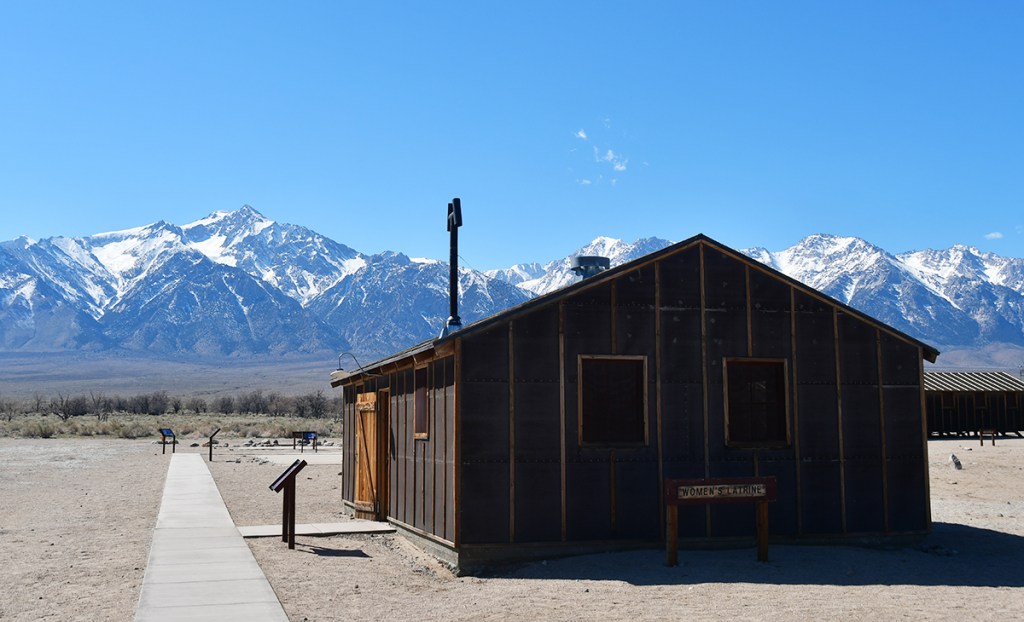 A historic wooden building labeled 'Women's Latrine' in a desert landscape with snow-capped mountains in the background under a clear blue sky.