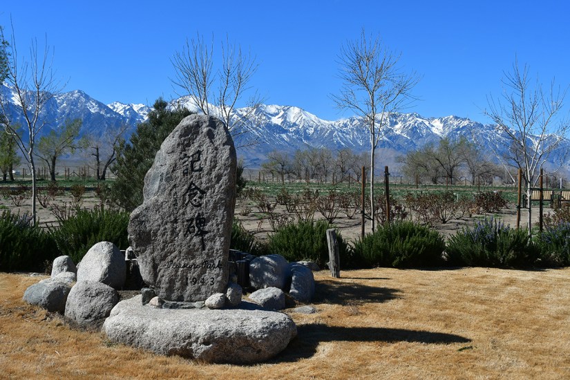 A memorial stone with inscriptions, surrounded by large rocks, in a landscape featuring snow-capped mountains and sparse vegetation under a clear blue sky.