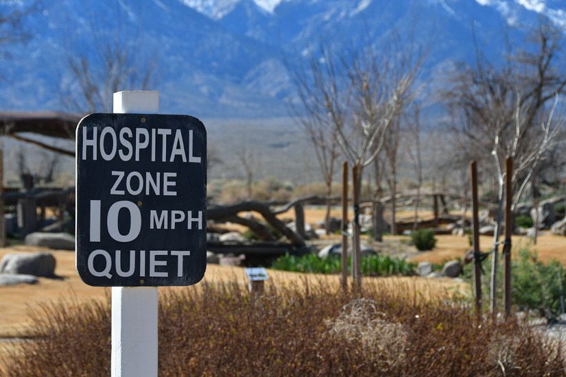 A sign indicating a hospital zone with a speed limit of 10 MPH and the word 'Quiet,' set against a backdrop of mountains and a landscaped area.