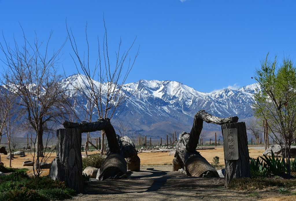 Scenic view of a rural pathway framed by wooden sculptures, leading towards snow-capped mountains under a clear blue sky.