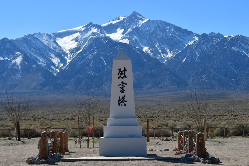 A tall monument with inscriptions stands in the foreground, framed by snow-capped mountains under a clear blue sky.