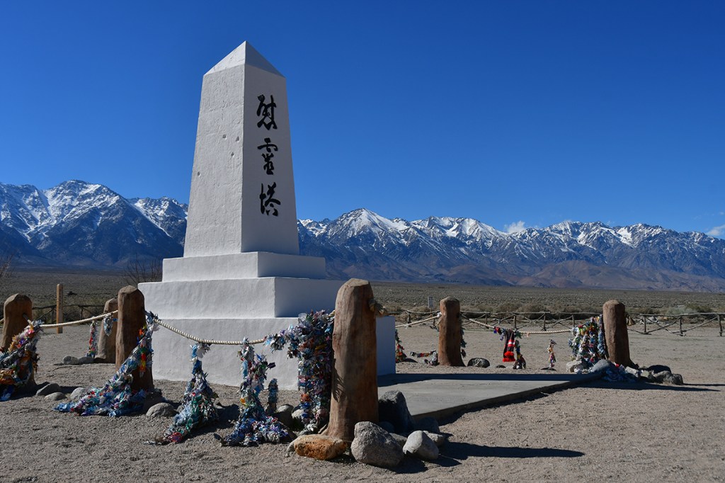 A white monument with inscriptions, surrounded by colorful decorations and rocks, set against a backdrop of snowy mountains and a clear blue sky.