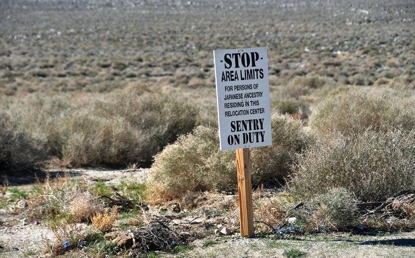 A sign stating 'STOP AREA LIMITS FOR PERSONS OF JAPANESE ANCESTRY RESIDING IN THIS RELOCATION CENTER SENTRY ON DUTY,' near a barren landscape with sparse vegetation.