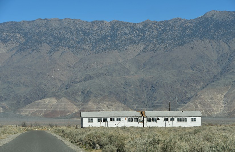 An abandoned white building with a flat roof in the foreground, set against a backdrop of rugged mountains and clear blue sky.