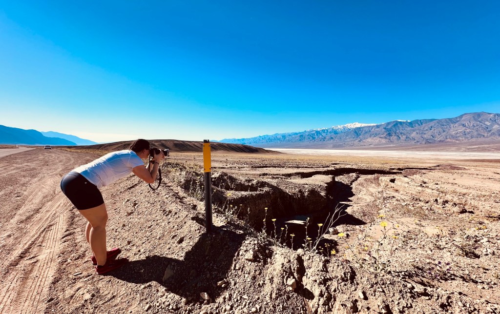 A photographer captures an image in a vast desert landscape with mountains in the background and a clear blue sky. A yellow marker stands near a exposed hole in the ground.