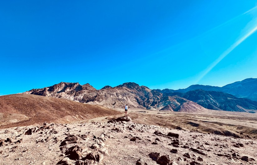 A lone hiker stands on a rock in a vast desert landscape with mountains in the background under a clear blue sky.