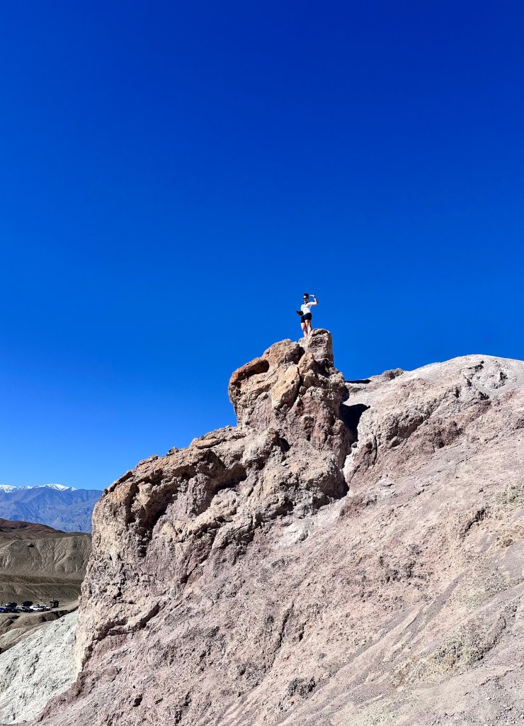 A person in a white shirt and shorts standing triumphantly on a rocky outcrop against a clear blue sky, with mountains visible in the background.