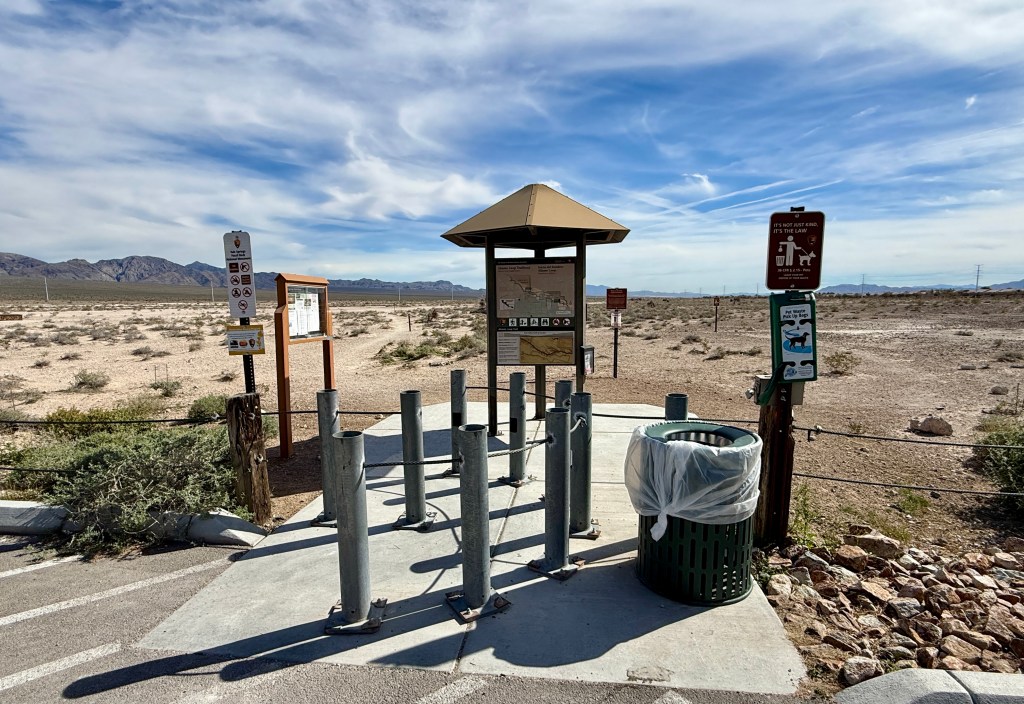 A trailhead area featuring informational signs, a covered map station, and a trash bin in a desert landscape.