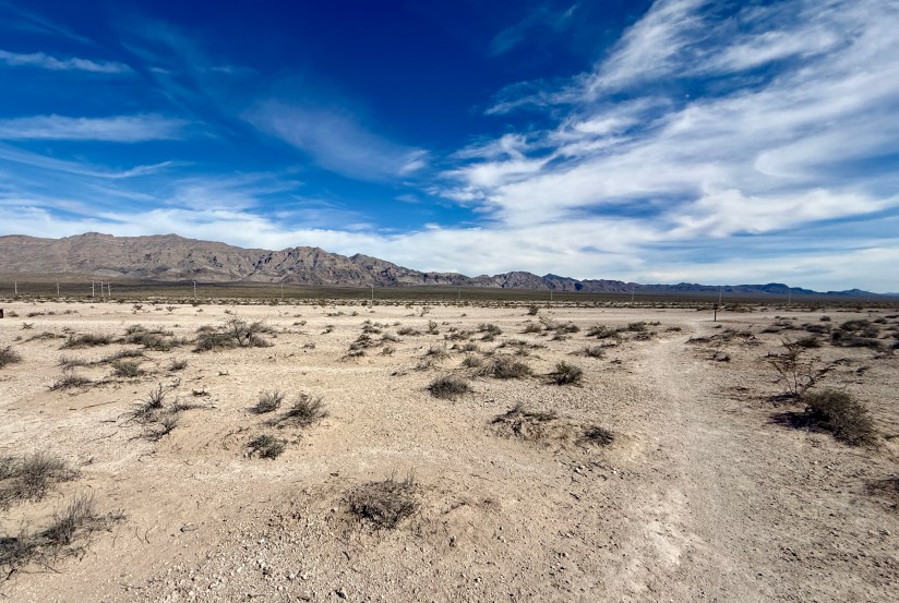 A barren desert landscape with sparse vegetation and a clear blue sky, featuring distant mountains under partly cloudy conditions.
