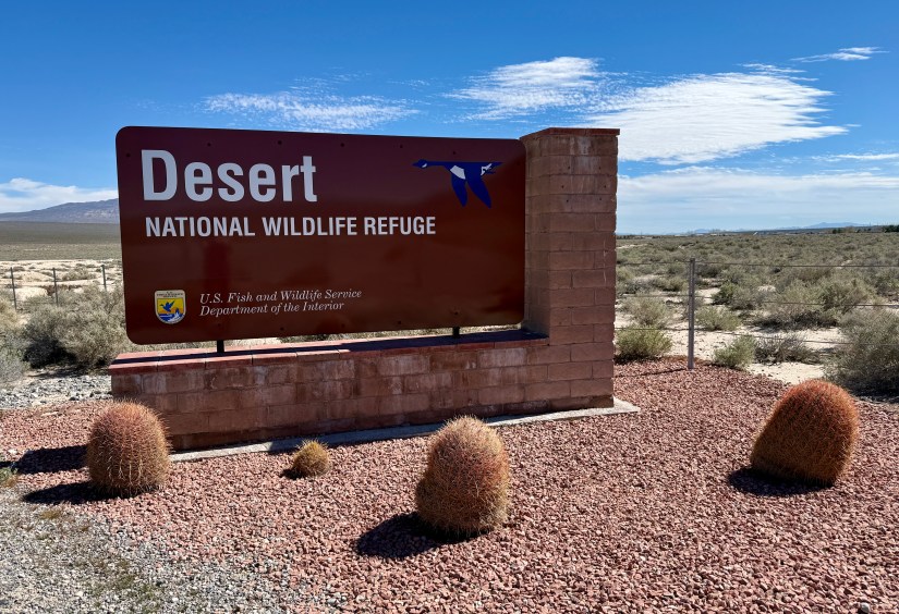 Sign for the Desert National Wildlife Refuge, featuring a red background with the text 'Desert NATIONAL WILDLIFE REFUGE' and a blue bird design, surrounded by desert landscape and cacti.