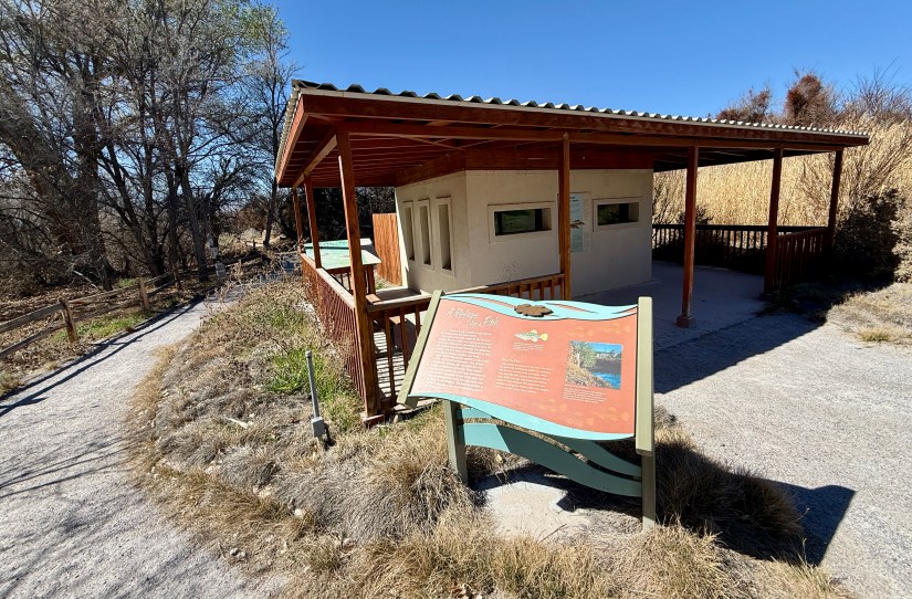 A wooden pavilion with informational signage in front, surrounded by a walking path and dry grass, set against a clear blue sky.