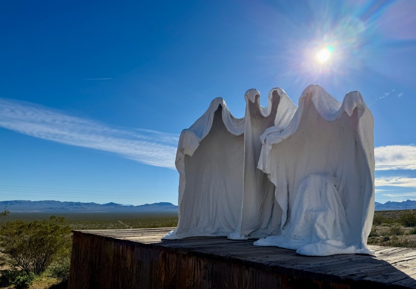 A white sculpture depicting three ghostly figures draped in flowing fabric, positioned on a wooden platform against a clear blue sky with sunlight.