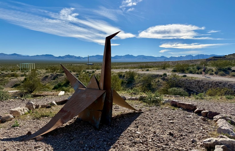 A large metal sculpture resembling a crane situated in a desert landscape with mountains in the distance under a clear blue sky.