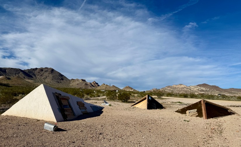 Three simple geometric structures, two wooden and one concrete, sit in a desert landscape with mountains in the background under a partly cloudy sky.