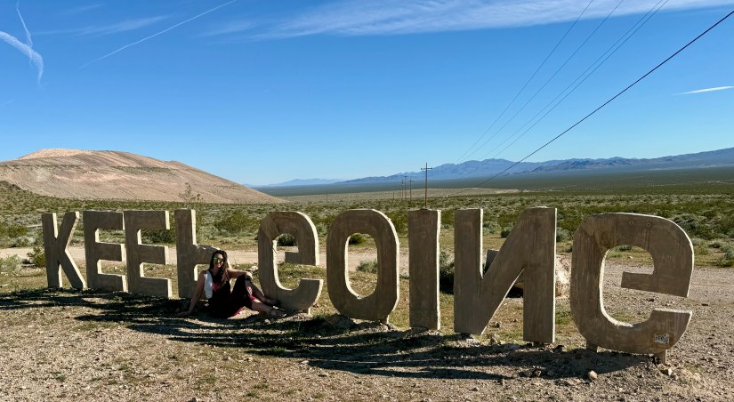 A person sitting next to large wooden letters spelling 'KEEP COINING' in a desert landscape with mountains in the background.