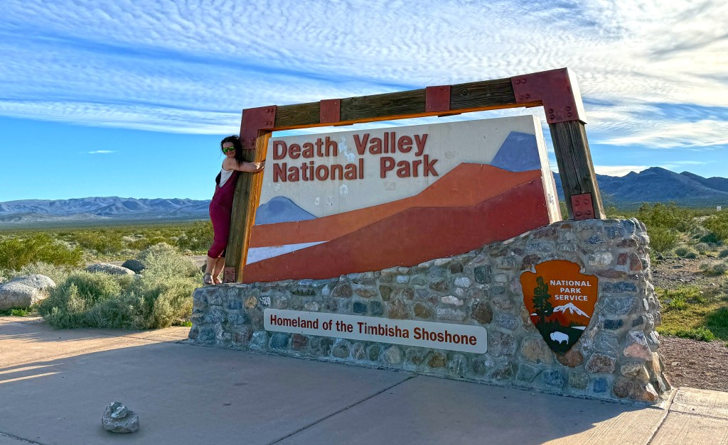 A visitor poses next to the Death Valley National Park sign, showcasing vibrant colors and a mountainous desert landscape in the background.