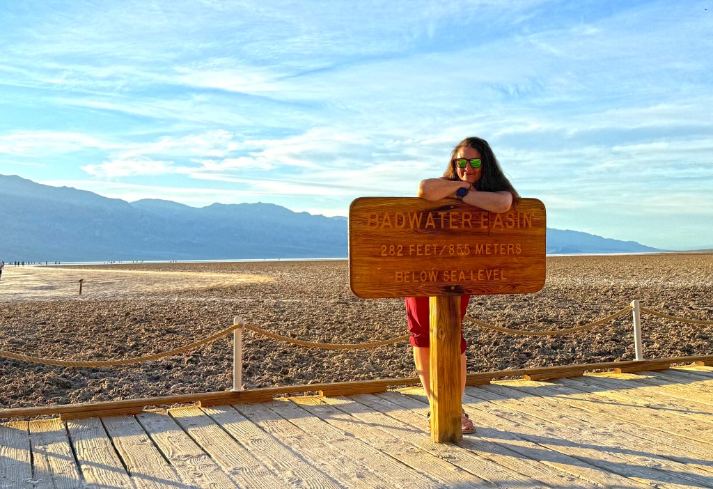 Person leaning against a sign for Badwater Basin, the lowest point in North America, with arid landscape and mountains in the background.