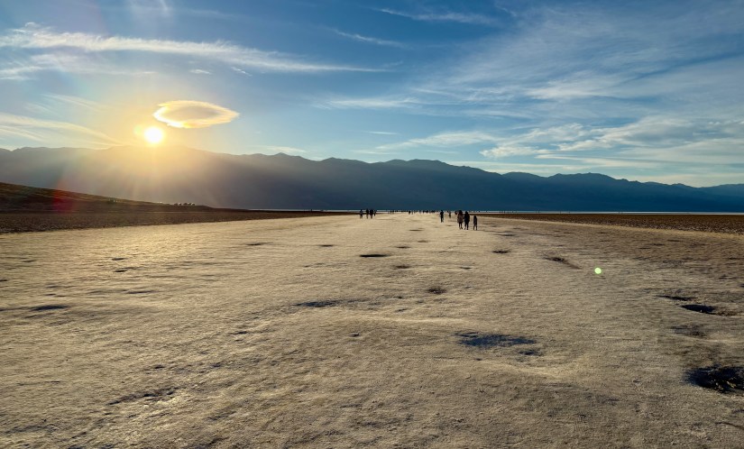 A serene landscape of a salt flat at sunset, with the sun hovering above distant mountains and people walking along a wide, dry pathway.