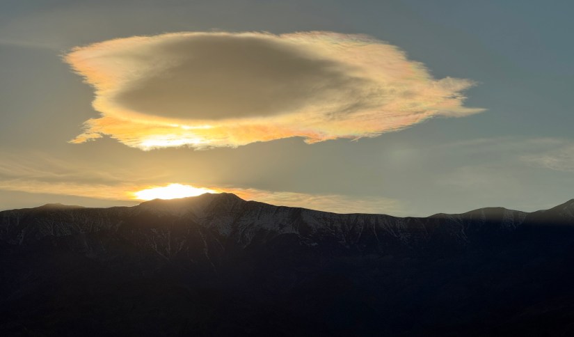 A dramatic sunset view behind mountains, featuring a large, circular cloud illuminated by warm colors from the setting sun.
