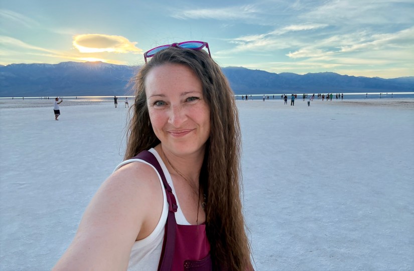 A smiling woman with long hair, wearing a white tank top and maroon overalls, stands in the foreground of a vast salt flat during sunset, with mountains and other visitors in the background.