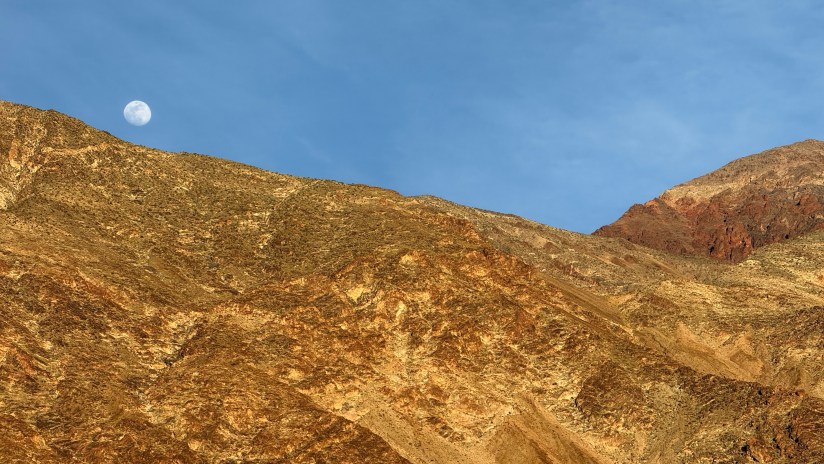 A panoramic view of rugged mountains with golden-brown slopes under a clear blue sky, featuring a bright full moon in the top left corner.