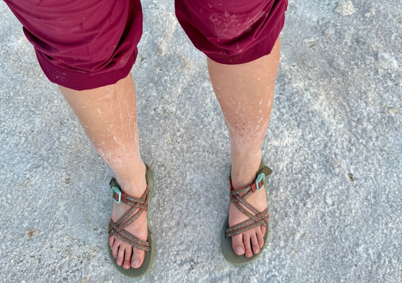 Close-up of a person's legs wearing maroon shorts and outdoor sandals, with visible salt residue on the skin, standing on a sandy surface.
