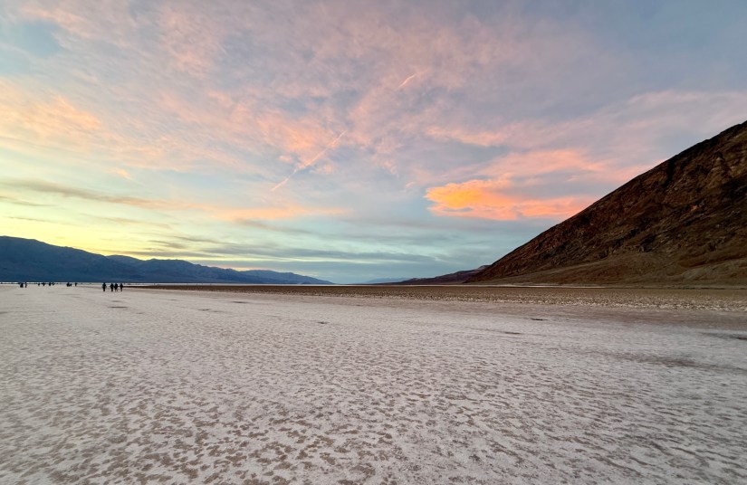 A vast, dry salt flat with a colorful sunset sky featuring shades of pink and blue, surrounded by distant mountains.