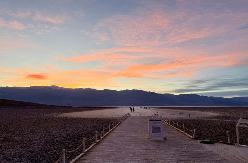 A wooden boardwalk leads into a scenic landscape at sunset, with colorful clouds above and distant mountains in the background. People are seen walking in the distance.