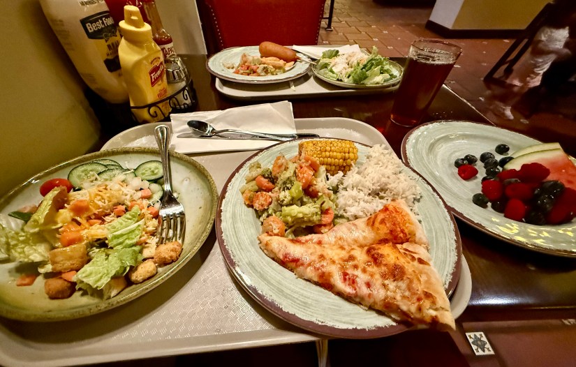 A tray with various food items including a salad with lettuce, tomatoes, and croutons, a main plate featuring a slice of pizza, shrimp stir-fry with vegetables, rice, and corn, alongside a dessert plate of watermelon and mixed berries. Condiments such as mustard and ketchup are visible.