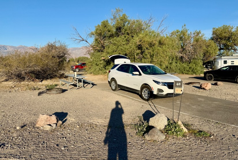 A campsite with a white SUV parked, a red vehicle in the background, and a picnic table nearby, surrounded by desert vegetation and rocks.