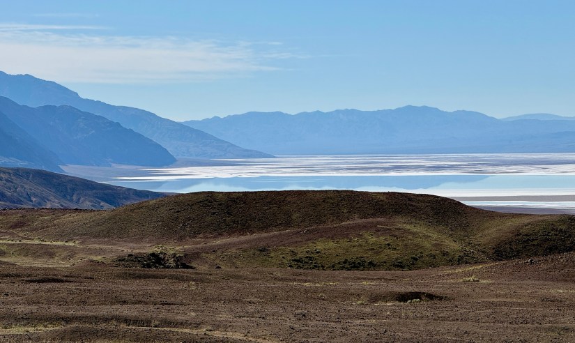 Scenic landscape of a dry desert area with mountains in the background and a reflective salt flat under a clear blue sky.