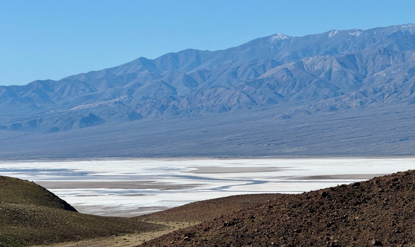 A panoramic view of a desert landscape featuring flat, white salt flats in the foreground and a range of rugged mountains in the background under a clear blue sky.